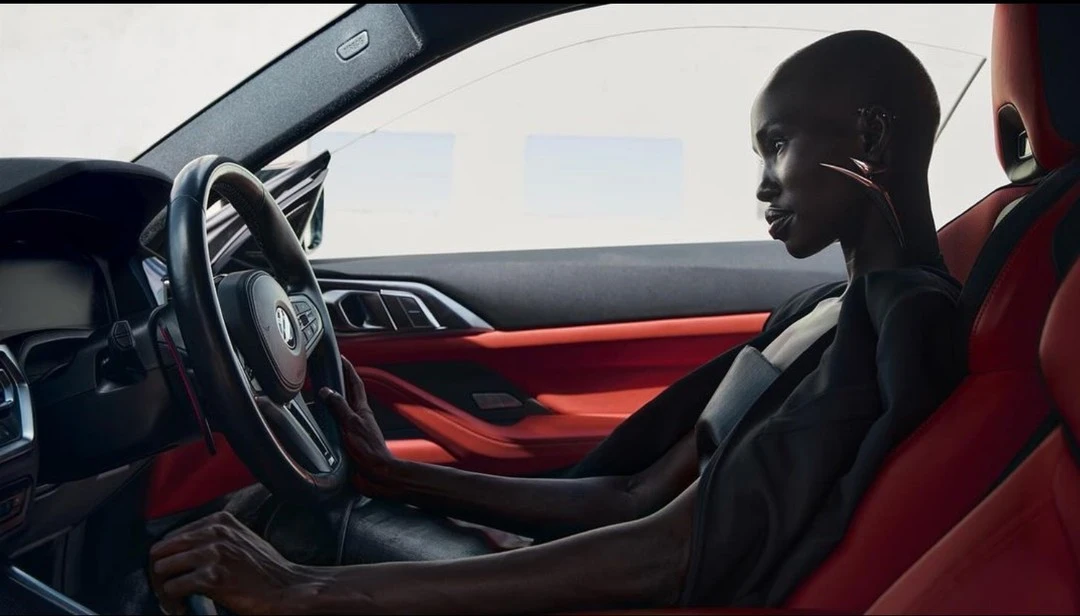 Adult woman sits inside a car with red interior, holding the steering wheel. Automotive fashion shot in natural lighting.