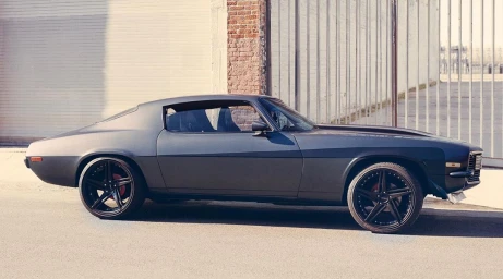 Profile view of a custom dark grey vintage muscle car parked on an asphalt street alongside a brick building exterior.
