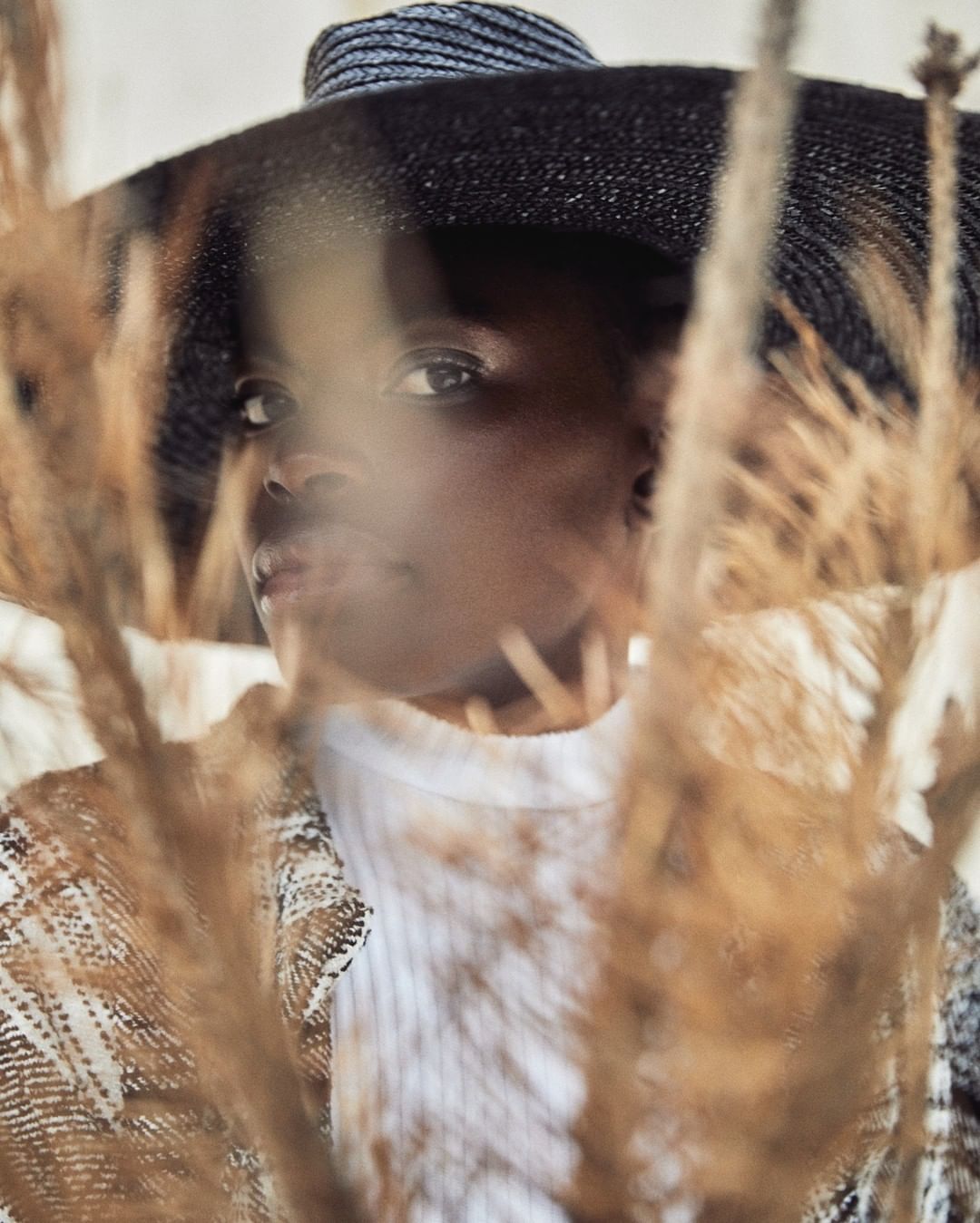 Woman in hat poses behind wheatgrass stalks in an eye-level indoor portrait with soft lighting. Fashion and beauty portrait.