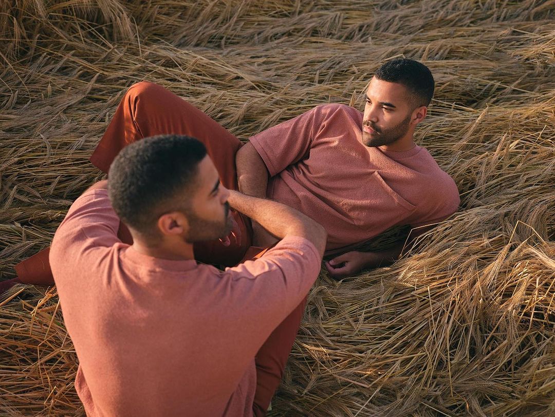 Two adult men laying in a field of dried wheat wearing rust-colored clothing, looking at each other in an outdoor setting.