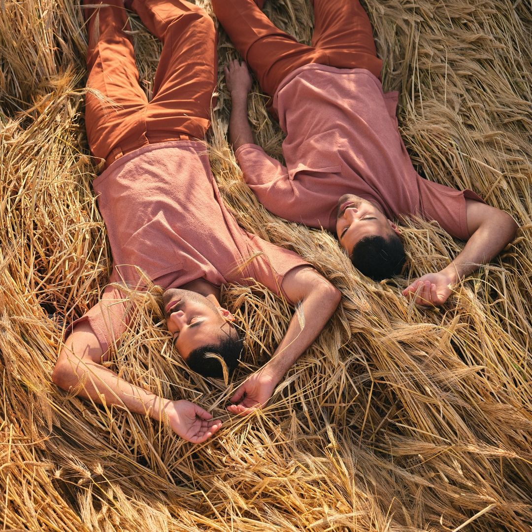 Two men in matching peach-colored shirts and terracotta pants lie in a field of golden wheat, capturing a peaceful and serene rural moment.