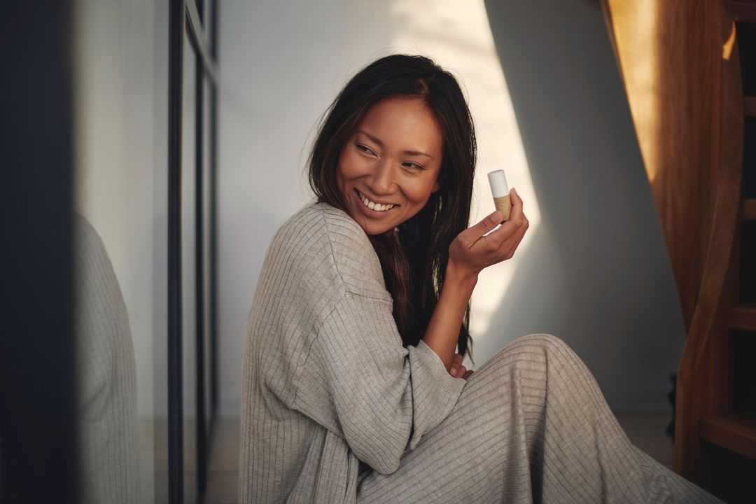 Smiling Asian woman in loungewear sitting on floor, holding small cosmetic product, near window with natural light, interior setting.