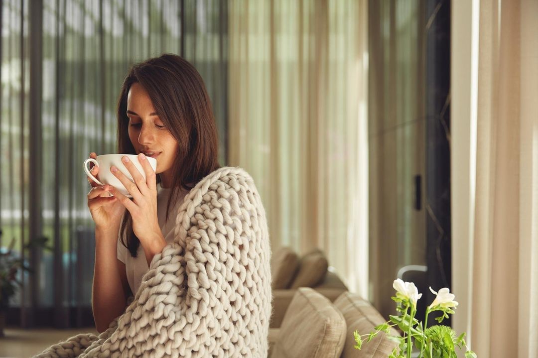 Woman drinking tea wrapped in chunky knit blanket sitting on couch at home. Serene and cozy scene in a bright indoor setting.