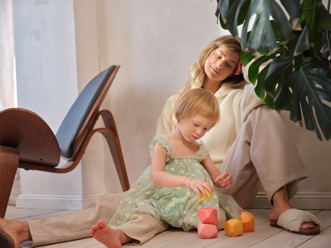 Young girl in green dress plays with wooden blocks beside her mother in a cream sweater and slippers inside a white room.