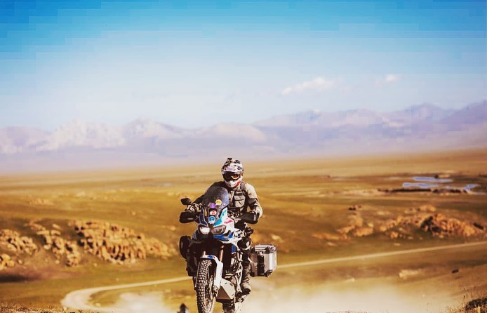 Adult male motorcycling on a dirt path through a grassy plain with distant mountains under a blue sky in the daytime.