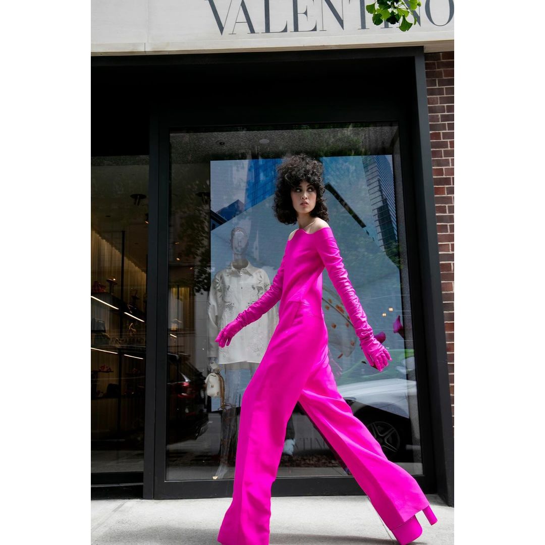 Elegant adult woman in a hot pink jumpsuit, leather gloves, and platform heels, walking past a designer boutique with a window display.