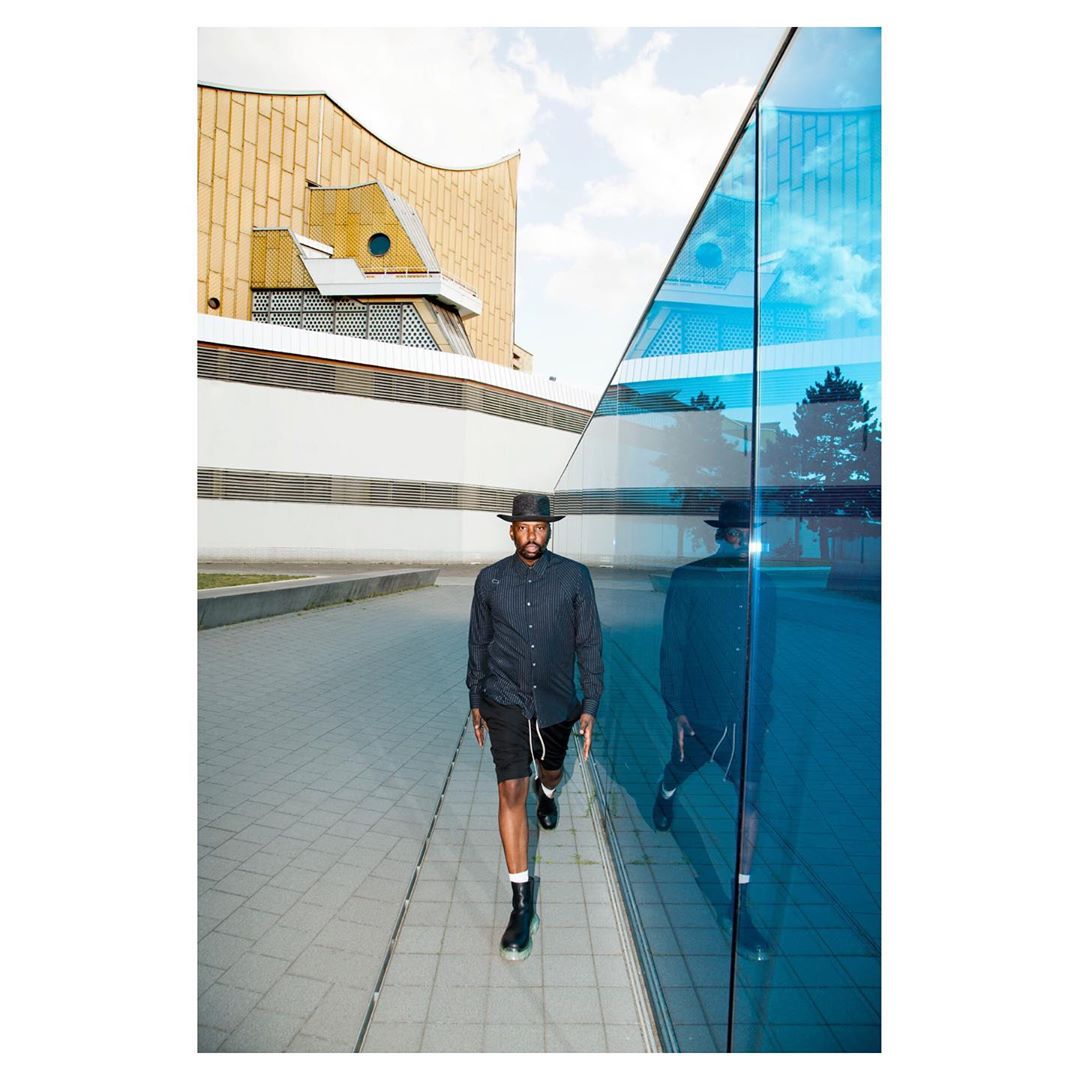 Man walks on a paved surface beside a modern building with reflective blue windows; he is wearing a hat, dark button shirt and shorts.