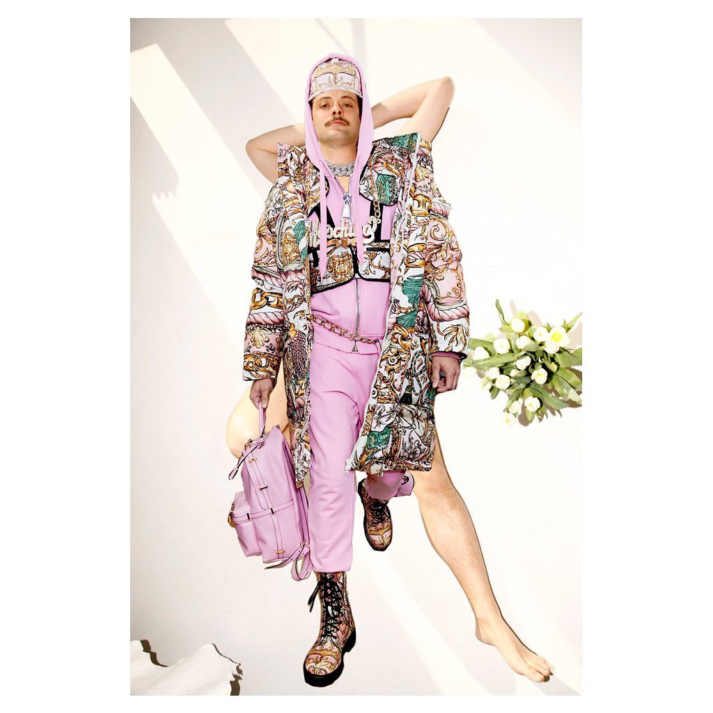 Man in a patterned coat and boots poses with a pink backpack, jacket, and pants against a neutral background in a fashion editorial shot.