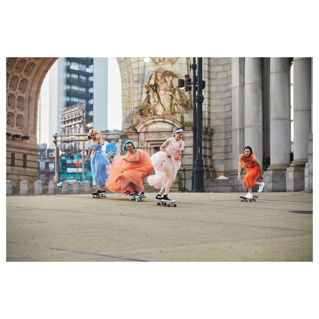 Four women in colorful dresses skateboard on a city bridge in New York City, with architecture and skyscrapers visible in the background.
