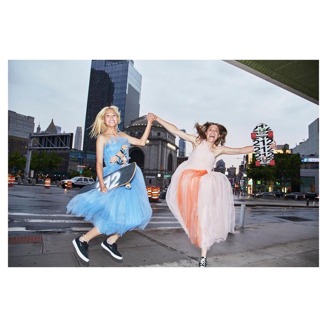 Two women in formal dresses hold skateboards and jump for joy on a wet city street with skyscrapers on a cloudy day.