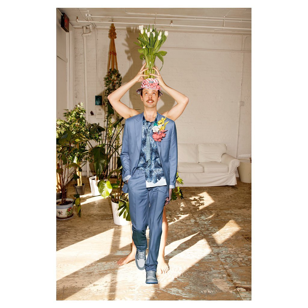 A man in a blue suit poses with tulips on his head, held by arms behind him, in a sunny studio with potted plants and hardwood floors.