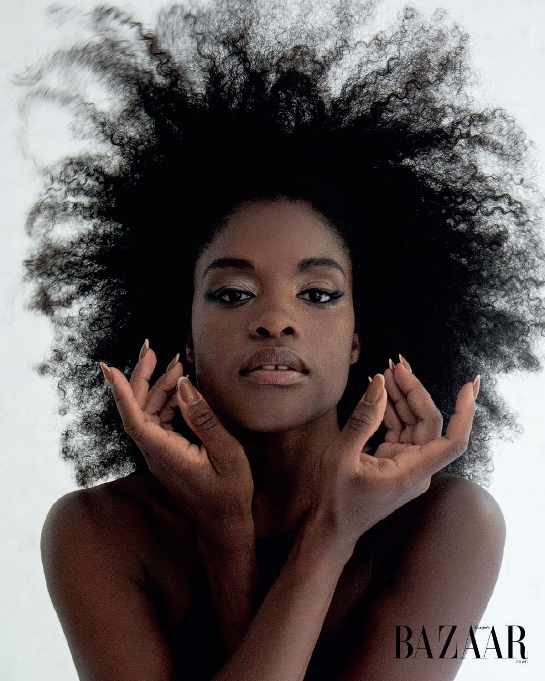Portrait of a woman with dark skin and an afro hairstyle. She is posing with her hands near her face against a light background.