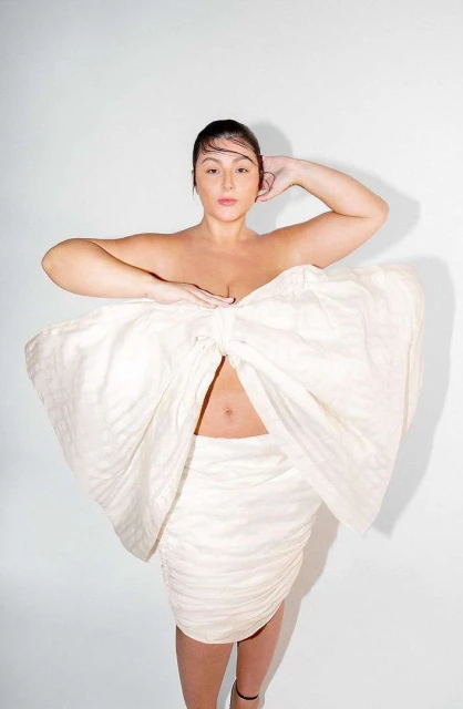 Woman posing in a large white bow dress with hands near her chest in a studio setting against a white backdrop, medium shot.