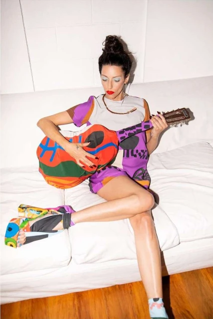 A woman with dark hair in a colorful outfit sits cross-legged playing an acoustic guitar on a white couch indoors, illuminated by bright lighting.