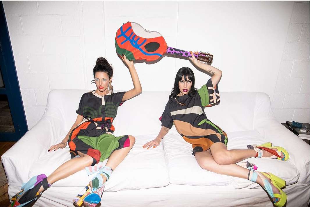 Two stylish adult women pose on a white couch in colorful outfits, holding a painted guitar-shaped object in an indoor setting.