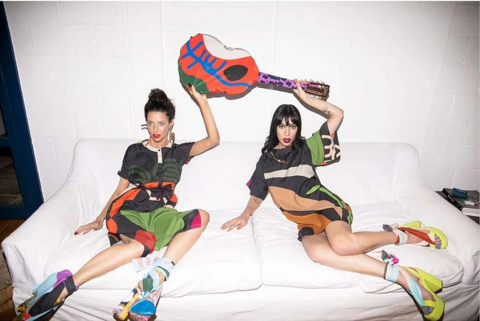 Two stylish adult women pose on a white couch in colorful outfits, holding a painted guitar-shaped object in an indoor setting.
