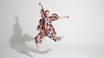 A woman in a colorful patterned outfit leaps barefoot against a white backdrop in a dynamic pose with arms raised overhead.