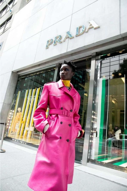 Woman in a pink Prada trench coat standing in front of a Prada store with a glass door and the Prada logo above the door.
