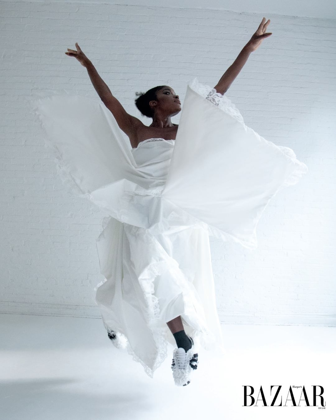 A ballerina in mid-air leaps in a white dress and sneakers against a white wall, with the 'BAZAAR' logo in the lower right corner.