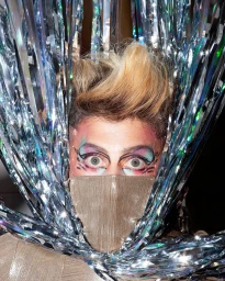 Close-up portrait by Marcelo Krasilcic of a person wearing elaborate eye makeup and a gold face mask framed by metallic tinsel streamers.
