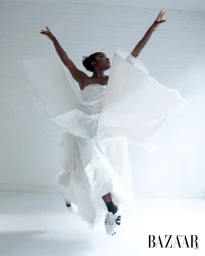 Black dancer leaping mid-air in a white ruffled dress with arms raised against a white brick wall by Marcelo Krasilcic