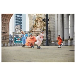 Four women in colourful dresses skateboarding together through an outdoor public square under an archway.