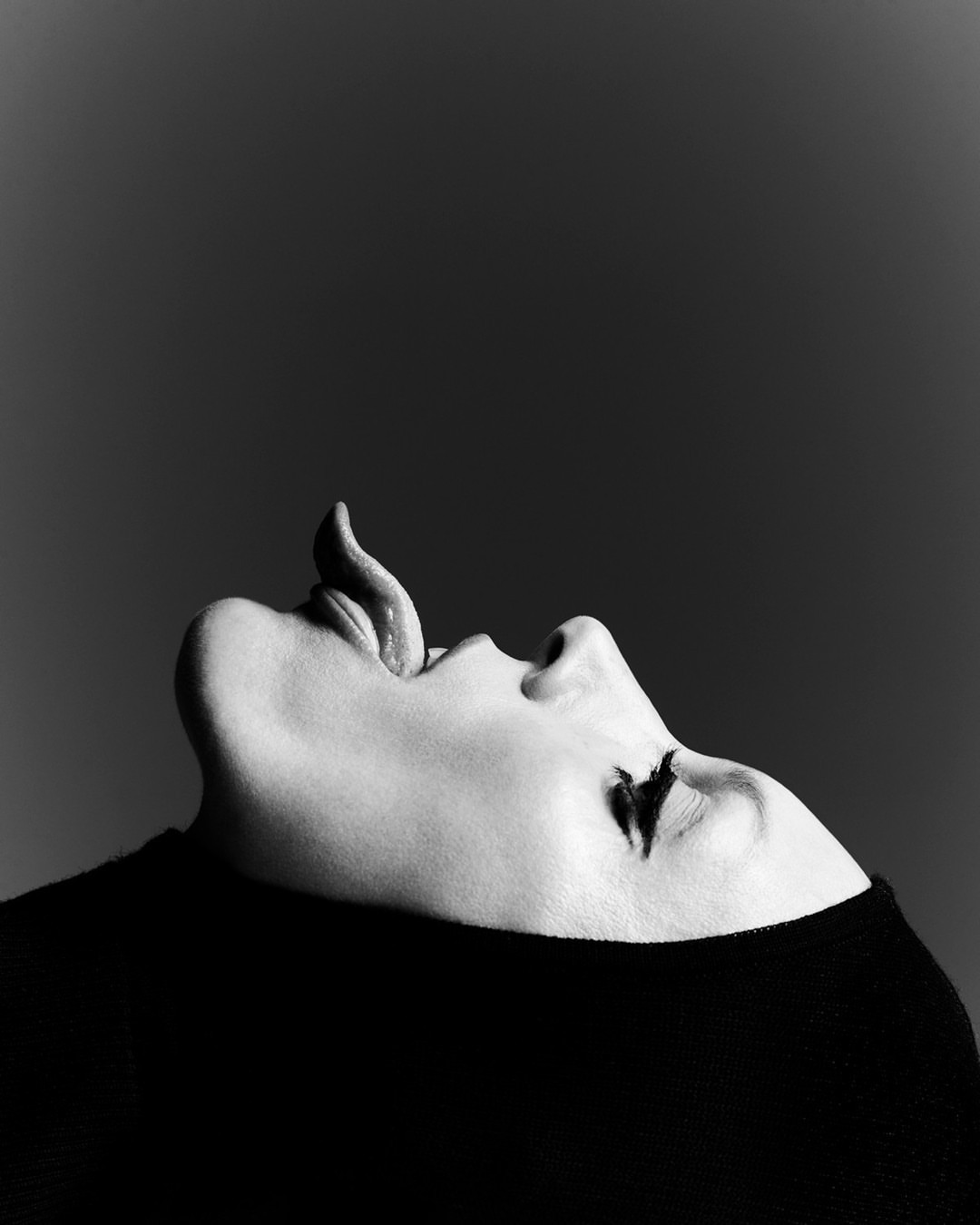 Close-up black and white portrait of a woman with her head tilted back and her tongue extended up, against a solid gray backdrop.