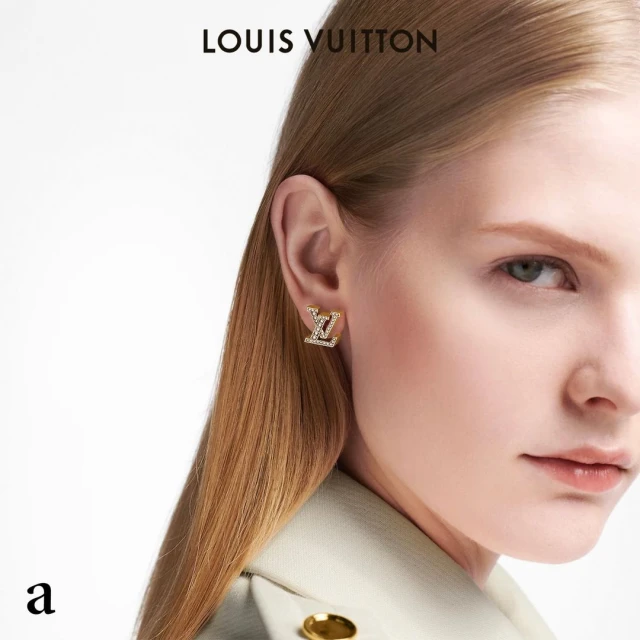 Blond woman's face in a close-up shot, showcasing a gold LV-logo earring, wearing a light-yellow blazer against a white backdrop.