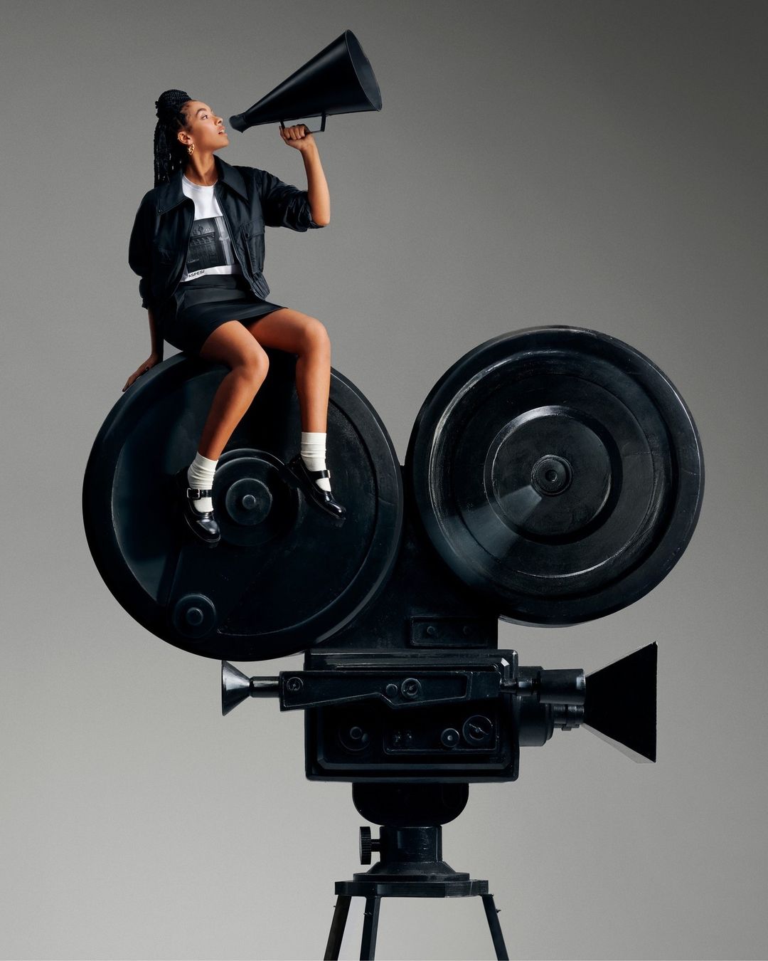 A young woman sits on a vintage camera, holding a megaphone against a gray backdrop, with a classic and cinematic feel.