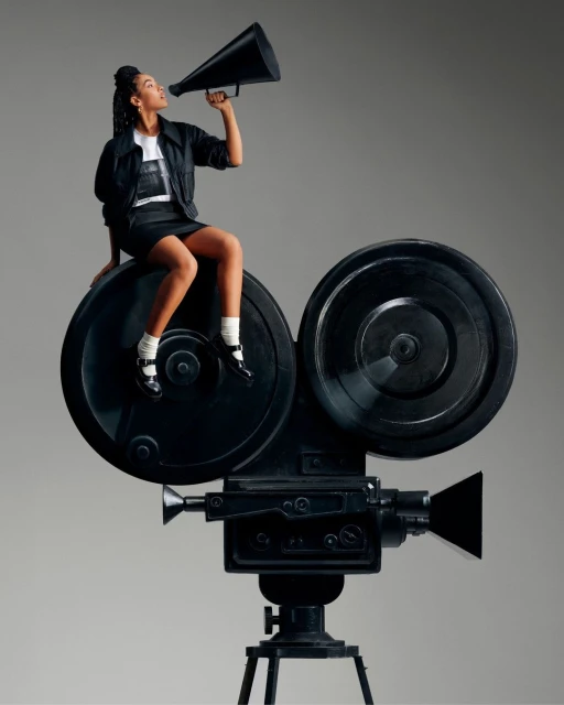 A young woman sits on a vintage camera, holding a megaphone against a gray backdrop, with a classic and cinematic feel.