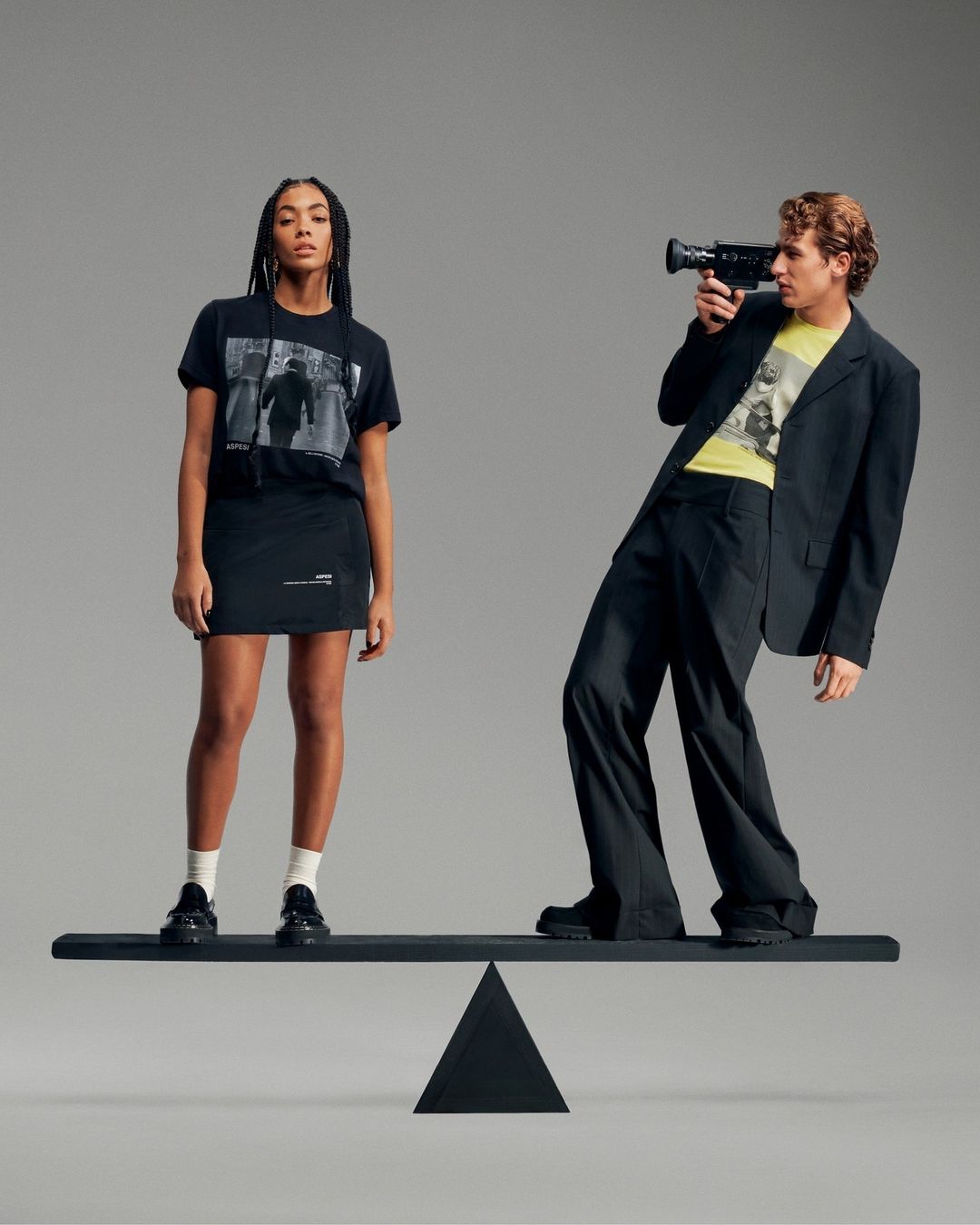 Two adult people, a woman and a man, stand on a black balance beam against a gray background in a fashion studio setting.