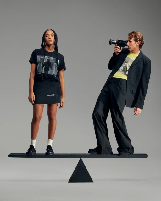 Two adult people, a woman and a man, stand on a black balance beam against a gray background in a fashion studio setting.