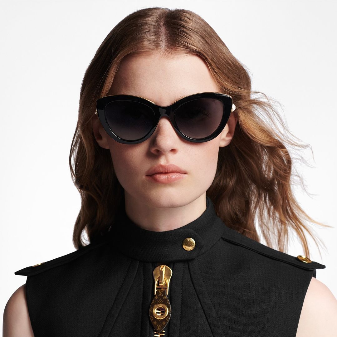 Close-up of a woman wearing sunglasses and a black sleeveless top with a zipper, against a white background in a studio setting.