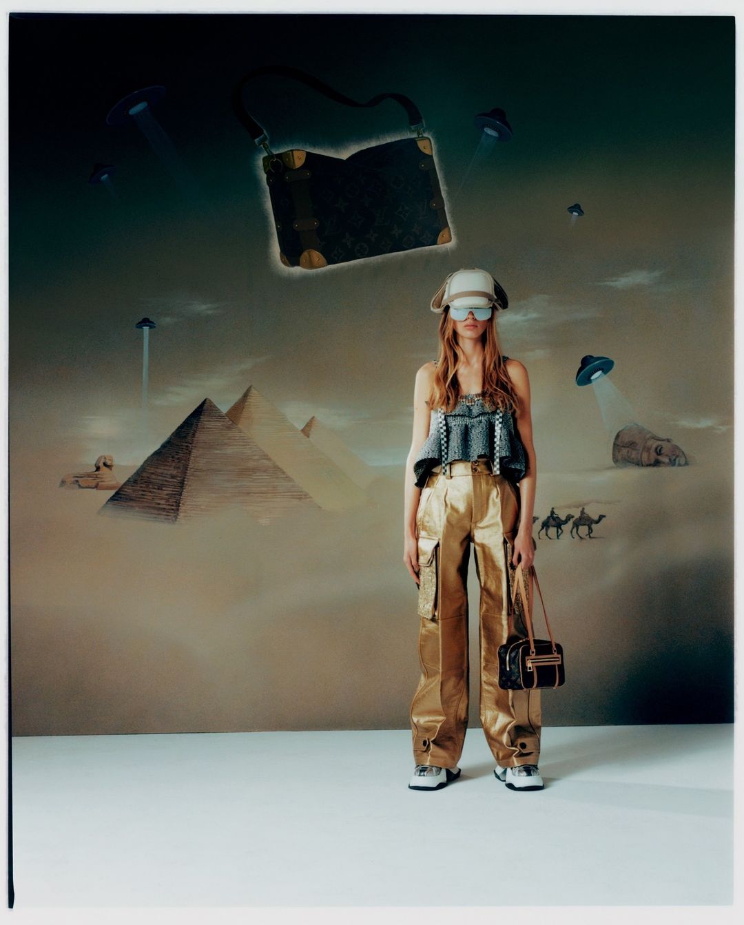 A woman in a hat and metallic pants poses with bags in front of a wall art backdrop with pyramids and flying saucers.