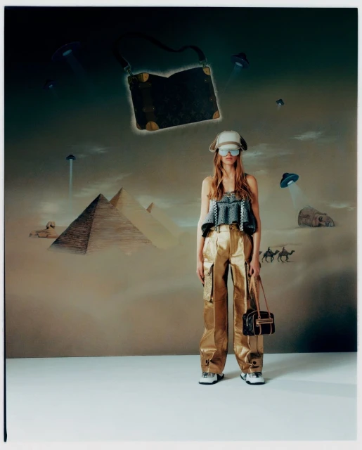 A woman in a hat and metallic pants poses with bags in front of a wall art backdrop with pyramids and flying saucers.