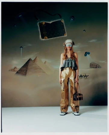 A woman in a hat and metallic pants poses with bags in front of a wall art backdrop with pyramids and flying saucers.