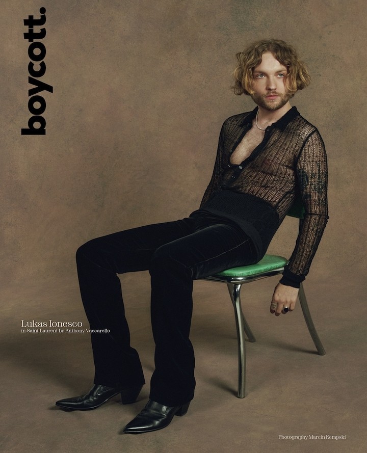 Blonde male model sits on green chair wearing a sheer black top, black velvet pants, and black leather shoes in a studio.
