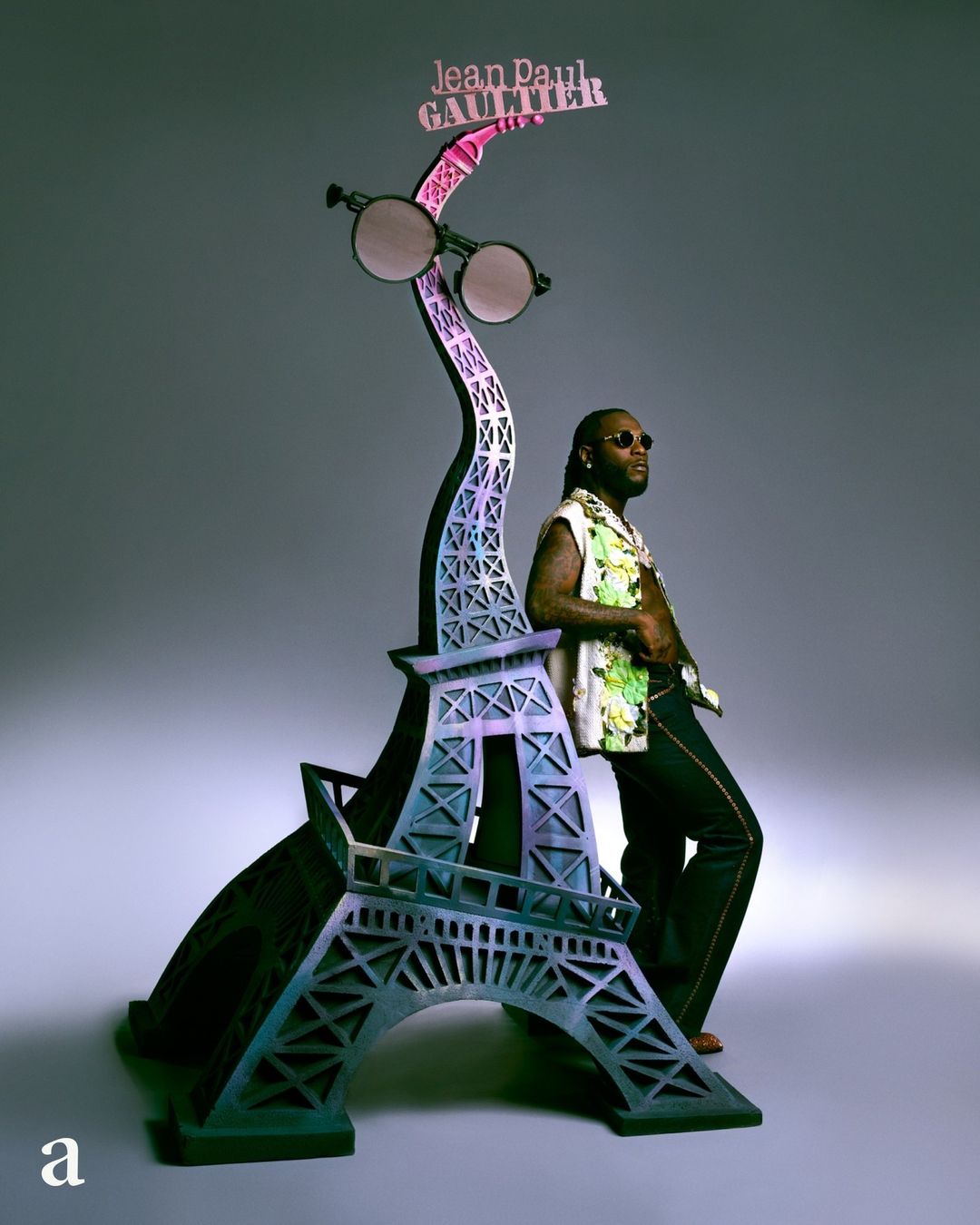 Adult male poses next to the Eiffel Tower display stand with round glasses and the brand name Jean Paul Gaultier on top.