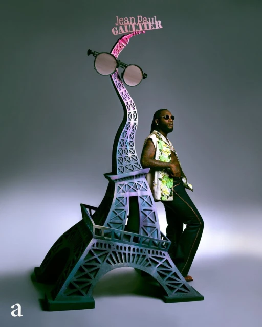 Adult male poses next to the Eiffel Tower display stand with round glasses and the brand name Jean Paul Gaultier on top.