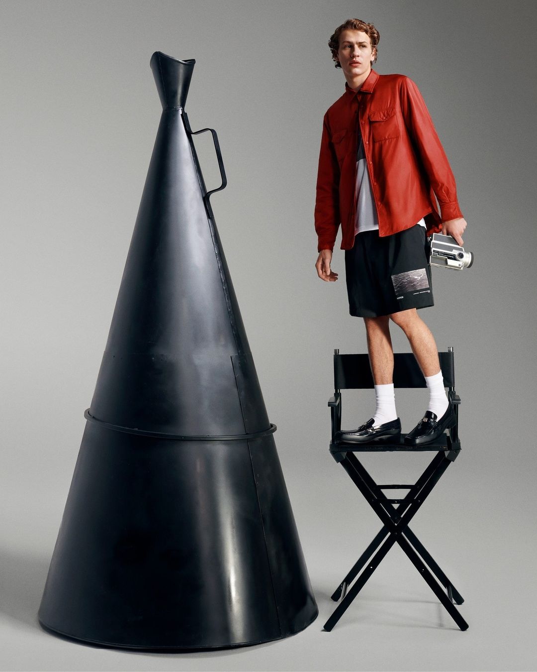 Young adult male poses on a black director's chair next to an oversized megaphone, wearing red shirt, black shorts, and holding a camera.