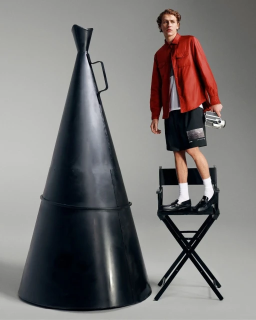 Young adult male poses on a black director's chair next to an oversized megaphone, wearing red shirt, black shorts, and holding a camera.