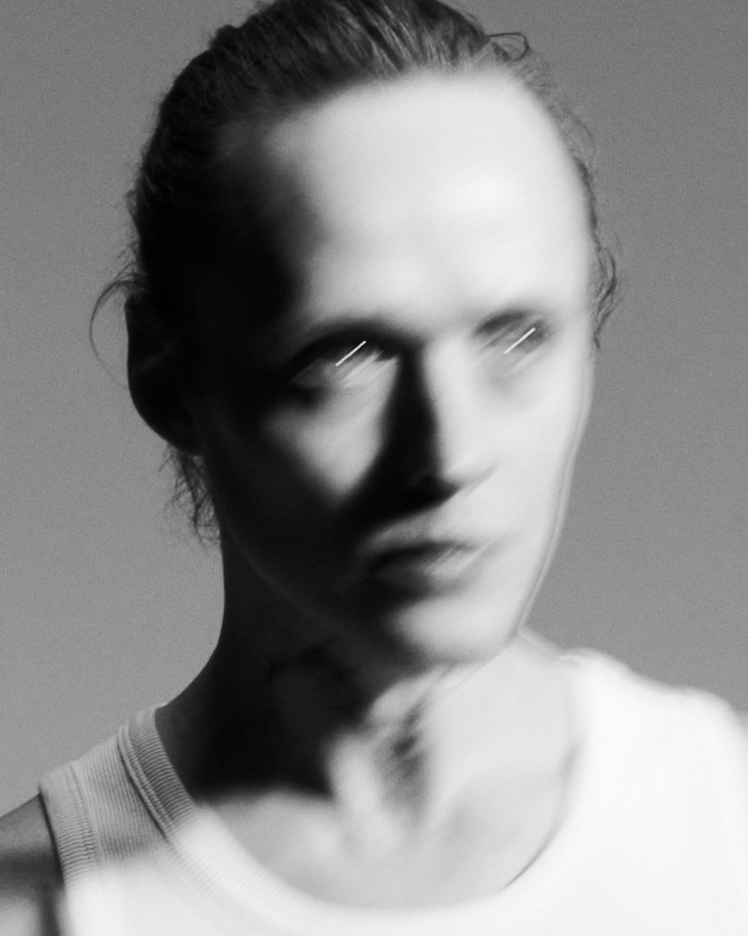 Monochrome portrait of a blurry man with a tank top, hair in a bun, and a choker necklace. The background is plain grey.