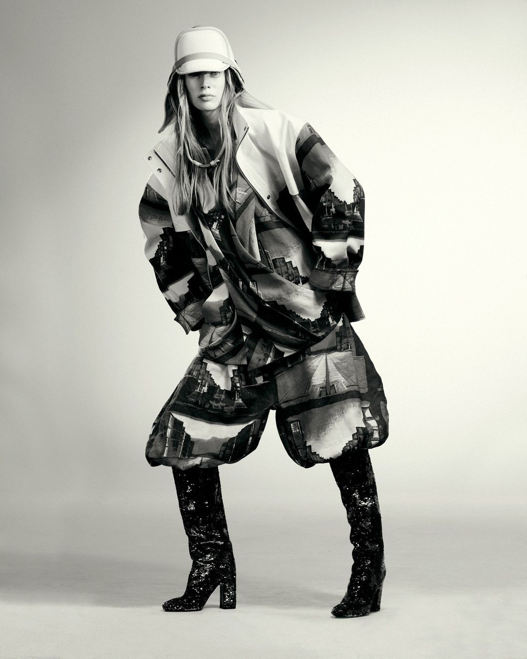 Woman poses in a fashion shot wearing a patterned jacket and shorts with shiny black boots against a neutral backdrop in a studio setting.