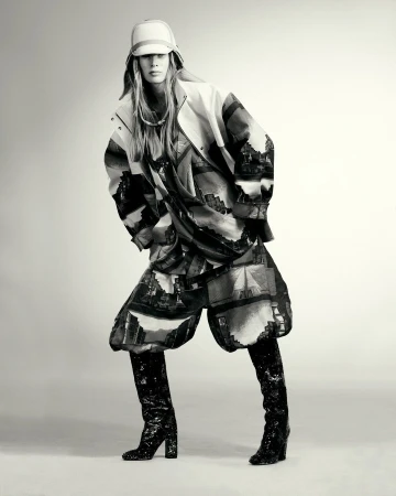Woman poses in a fashion shot wearing a patterned jacket and shorts with shiny black boots against a neutral backdrop in a studio setting.