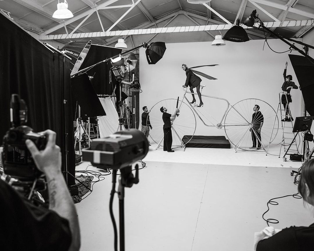 In a photo studio, a fashion shoot with a model on a large bicycle prop is captured by a team using professional lighting equipment.