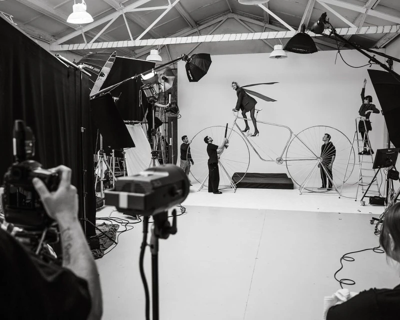 In a photo studio, a fashion shoot with a model on a large bicycle prop is captured by a team using professional lighting equipment.