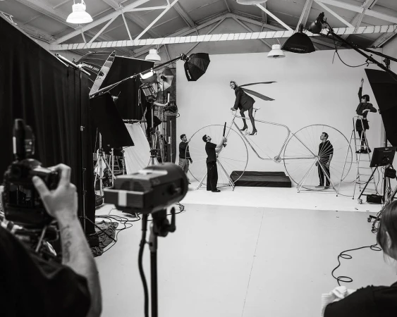 In a photo studio, a fashion shoot with a model on a large bicycle prop is captured by a team using professional lighting equipment.
