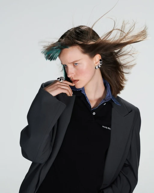 Portrait of a young adult female fashion model with dark hair and green highlights wearing a blazer, polo shirt, and statement jewelry.
