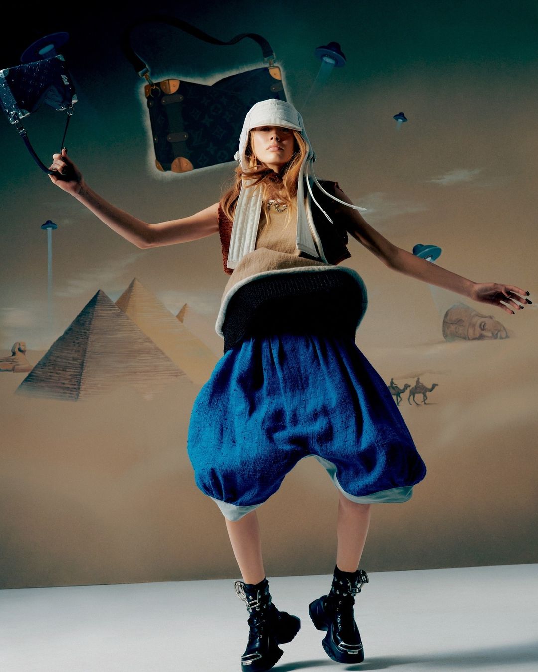 Fashion editorial shot of a woman in blue shorts, boots, and a hat, holding a purse, with pyramids and UFOs in the background.
