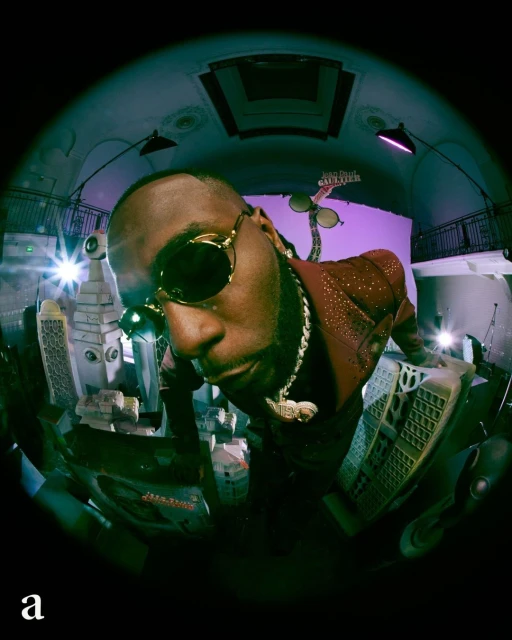 Burna Boy captured with a fisheye lens in a surreal setting. The artist wears sunglasses and a sparkling jacket, with cityscapes in the frame.